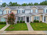 Two-story townhouse at 1567 Quail Circle with gray siding, black front door, and white trim, surrounded by a green lawn and bushes. A small tree is planted in front; sidewalks run parallel to the building and street. Located in Creedmoor, this home is attached to neighboring townhouses.
