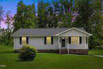 Charming single-story white house with dark shutters, a covered front porch, and a green front door awaits you at 205 Polly Drive, Oxford. The home is surrounded by lush grass with trees in the background, while the sky above is partly pink and blue, suggesting a peaceful sunrise or sunset.