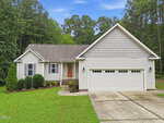 Charming single-story beige house at 85 Copper Creek Drive, Youngsville, featuring a welcoming front porch, gabled roof, two-car garage, and a concrete driveway. Beautiful bushes and a manicured lawn grace the front yard, all surrounded by tall trees and set beneath a partly cloudy sky.