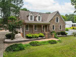 Nestled at 8429 Wycombe Ridge Way in Wake Forest, this two-story house features a charming stone facade, three dormer windows, a covered front porch with columns, and a beautifully landscaped front yard. The concrete driveway leads to the home, while mature trees and neighboring houses are visible in the background under a partly cloudy sky.