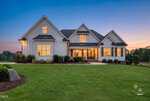 A modern two-story house with white siding, large front windows, and a covered porch sits on a well-manicured lawn at sunset in Zebulon. Located at 45 Water Willow Lane, outdoor lights are on, and landscaping includes shrubs and small trees.
