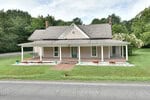 Located at 102 Creedmoor Road in Stem, this charming single-story farmhouse features a full-width covered porch, gray siding, and two brick chimneys. The home boasts a teal front door, potted plants, and a green lawn bordered by trees.