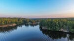 A calm lake near 111 Osage Drive, Louisburg is surrounded by dense green trees under a clear blue sky at sunset. Several docks and small boats are visible along the shoreline, while the water beautifully reflects the trees and sky.