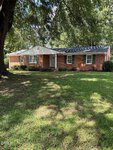Charming single-story red brick house with white trim and green shutters at 201 Forest Road in Oxford. The white front door welcomes you from a green lawn lined with bushes, while large trees provide shade over the yard and house.
