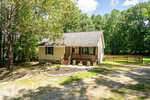 A small, single-story beige house with a covered front porch, steps, and potted plants at 210 Deer Run Drive. Surrounded by trees in the peaceful Franklinton area, it features a gravel driveway and a wooden fence in the background. The sky is partly cloudy.