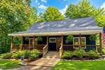 A charming blue house at 108 Cedar Creek Lane in Youngsville features a large front porch with wooden railings, hanging plants, and an American flag, all surrounded by green shrubs and trees under a sunny sky. Steps lead up to the inviting front door.