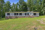 Single-story manufactured home with beige siding and a brick foundation, set on a grassy lawn with scattered trees and a wooded background in Franklinton. A small set of steps leads to the front door at 245 Oak Forest Lane. The sky is clear and sunny.