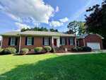 Charming single-story brick house located at 66 Pine View Road in Henderson, featuring classic black shutters, a striking red front door, inviting front steps, and an attached two-car garage. Enjoy a well-kept lawn with trimmed bushes and mature trees in the background under a partly cloudy sky.