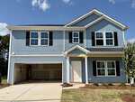 A charming two-story blue house with white trim, black shutters, and an attached two-car garage sits at 180 Buckthorn Drive in Youngsville. The garage door is open, revealing ample space, while a small covered front porch welcomes visitors. The yard features patches of grass and neatly arranged small shrubs.