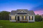 A small white house with a covered front porch, black shutters, and a gabled roof sits on a grassy lawn at 20 Bambi Lane in Louisburg. Trees line the background under a blue and purple sunset sky. Lights are on inside the house.