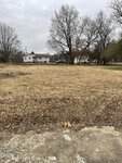 A vacant lot with dry grass and a few scattered leaves in the foreground. Bare trees line the back, and houses and a parked car are visible across the street under a cloudy sky in Oxford, near 513 Hillsboro Street.