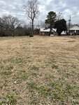 A grassy vacant lot with sparse green and brown patches, bordered by a road on 507 Hillsboro Street in Oxford. Leafless trees and two houses are visible in the background under a cloudy sky.