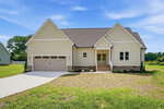 Single-story beige house with a dark shingle roof, attached two-car garage, front steps leading to a covered entry, and a concrete driveway. Nestled on 35 Cotton Drive in Louisburg, the home features a grass yard surrounding the property with trees in the background under a clear blue sky.