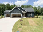 A new, dark blue single-story house with white trim, a three-car garage, and a freshly sodded lawn sits on a curved driveway at 65 Kettle Creek Drive in Zebulon. Trees and greenery are visible in the background under a partly cloudy sky.