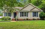 Charming single-story house at 114 Black Cloud Drive, Louisburg, featuring beige siding, blue shutters, and a classic gable roof. There are three front windows, a front door with steps and railing, a patch of lawn, and mature trees surrounding the property.