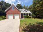 A charming red-brick house at 4336 N Greg Allen Way, Oxford, features two white garage doors, a covered front porch, and elegant dormer windows. The home boasts a wide driveway, lush green lawn, and is surrounded by trees under a blue sky with clouds.