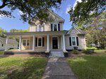 Two-story beige house with white trim, a covered front porch supported by columns, and a gabled roof. Located at 512 Raleigh Road in Oxford, this charming home features a concrete walkway leading to the steps and trees surrounding the yard. Blue sky with scattered clouds above perfectly frames the inviting setting.