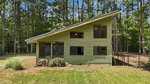 A green, modern house with a slanted roof sits surrounded by tall trees at 221 Allens Way. Large windows cover the front of the Kittrell home, and a small pile of wood is stacked near the foundation. There is a fenced area to the right.
