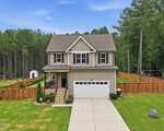 Charming two-story beige house at 265 Sorrel Drive in Franklinton, featuring a covered front porch, attached two-car garage, and fenced backyard. Surrounded by trees and a landscaped lawn, with a concrete driveway leading to the street.