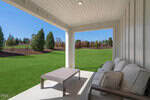 A covered patio at 346 Moose Meadow Way in Youngsville features a beige outdoor sofa and a rectangular coffee table, overlooking a green lawn and landscaped garden with trees and shrubs under a clear blue sky.