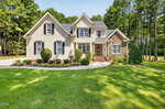 Two-story house at 135 River Club Way, Youngsville, featuring light-colored siding, stone accents on one side, symmetrical front windows, and a covered front porch. The home is surrounded by a manicured lawn, shrubs, and tall trees in the background.