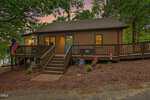 Charming brown single-story house at 207 Sagamore Drive, Louisburg, featuring a wooden deck and steps leading to a teal front door. Various flags are proudly displayed, and the property is surrounded by mature trees, with a lake partially visible in the background at sunset.