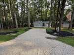 A gravel driveway leads to a small, light blue house surrounded by tall trees at 113 Kansas Drive in Louisburg. There are mulched garden beds on each side, a flag on the right, and a shed visible in the background.