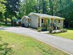 A single-story beige house at 1011 Shirley Drive, Henderson, features a covered front porch with black railings and a sloped driveway leading to a small shed in the backyard. The yard showcases green grass and flower beds, while tall trees surround the property.