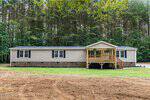 A beige manufactured home with black shutters and a brick foundation sits in front of a pine forest at 25 Green Pastures Road, Louisburg. The home features a covered wooden porch with railings and steps, and the yard is mostly bare dirt with some grass.