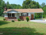 Charming single-story brick house on 3155 Kelly Road, Henderson, featuring a covered front porch, carport, and chimney. The yard boasts green grass and small shrubs, with trees and woods providing a scenic backdrop.