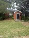 A small, one-story red brick house sits at 202 Mitchell Avenue in Franklinton, featuring a black front door, two windows, and a gabled roof. The yard is grassy with some patches of dirt, while bushes and trees surround the house.