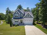 Charming two-story gray house with white trim at 135 Anna Marie Way, Youngsville. Featuring a covered front porch, attached two-car garage, and a neatly mowed lawn, this home is surrounded by trees and a clear blue sky, with a wide concrete driveway leading up to the residence.