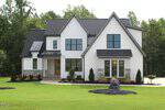 A modern two-story white house with black trim at 40 Serenity Woods Trail, Franklinton, features multiple peaked roofs, large windows, stone accents, and neat landscaping including bushes and mulched areas on a green lawn, set against a background of trees.