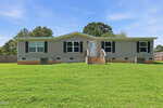 A beige, single-story manufactured home with black shutters and a small front porch sits on a grassy lawn at 6 A F Moore Road in Spring Hope, with trees and a wooden fence in the background under a blue sky.