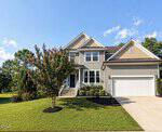 Two-story suburban house with beige siding, white trim, and a double garage located at 25 Summit Point in Youngsville. The front yard has neatly trimmed grass, landscaped bushes, a flowering tree, and a concrete driveway. The sky is blue with a few clouds.