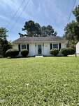 Small white single-story house with dark shutters, black roof, central front door, and trimmed bushes. Located at 240 Vicksboro Road in Henderson, this home sits on a well-maintained green lawn with trees in the background and a clear sky above.