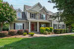 Charming two-story suburban house in Wake Forest featuring gray siding, stone accents, and crisp white trim. The inviting front porch at 380 Barn Hill Lane has elegant columns, while the landscaping showcases neatly mowed lawns, mature trees, and well-kept bushes. Multiple windows fill the home with natural light, and a bold red front door adds a welcoming touch.