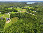 Aerial view of a rural neighborhood with scattered houses, winding roads, large green lawns, and dense forest areas. A lake is visible in the distance under a cloudy sky.