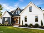 A modern farmhouse-style house at 25 Uptown Court in Zebulon, featuring white and dark gray vertical siding, large windows, a covered front porch, landscaped bushes, and a green lawn under a blue sky.