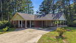 A one-story brick house with a gable roof, covered porch, and attached carport is nestled at 69 Dyking Road in Louisburg. The driveway curves toward the carport, while grass, shrubs, and tall pine trees surround the property.