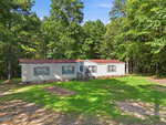 A single-story manufactured home with a brown roof and light gray siding sits on a grassy lot bordered by tall trees under a clear blue sky at 77 Baytree Lane, Henderson. A small wooden porch leads to the front door.