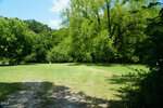 Open grassy area bordered by dense green trees under a blue sky with scattered clouds. Sunlight creates a mix of shadows and bright patches on the ground. A wooden picnic table is visible on the left side near the tree line, making this peaceful spot along 0-0 Halifax Road, Louisburg, an inviting place to relax outdoors.