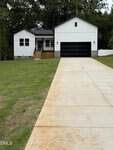 A white, single-story house with black trim and a black garage door sits at the end of a long concrete driveway at 244 Sequoia Drive in Louisburg. The lawn is spacious and green, with trees in the background and a small porch at the front entrance.