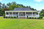 A single-story house at 102 Bartholomew Road in Louisburg features a wide front porch, white railings, gray siding, shuttered windows, and a sloped roof, surrounded by a well-kept lawn and trees in the background under a partly cloudy sky.