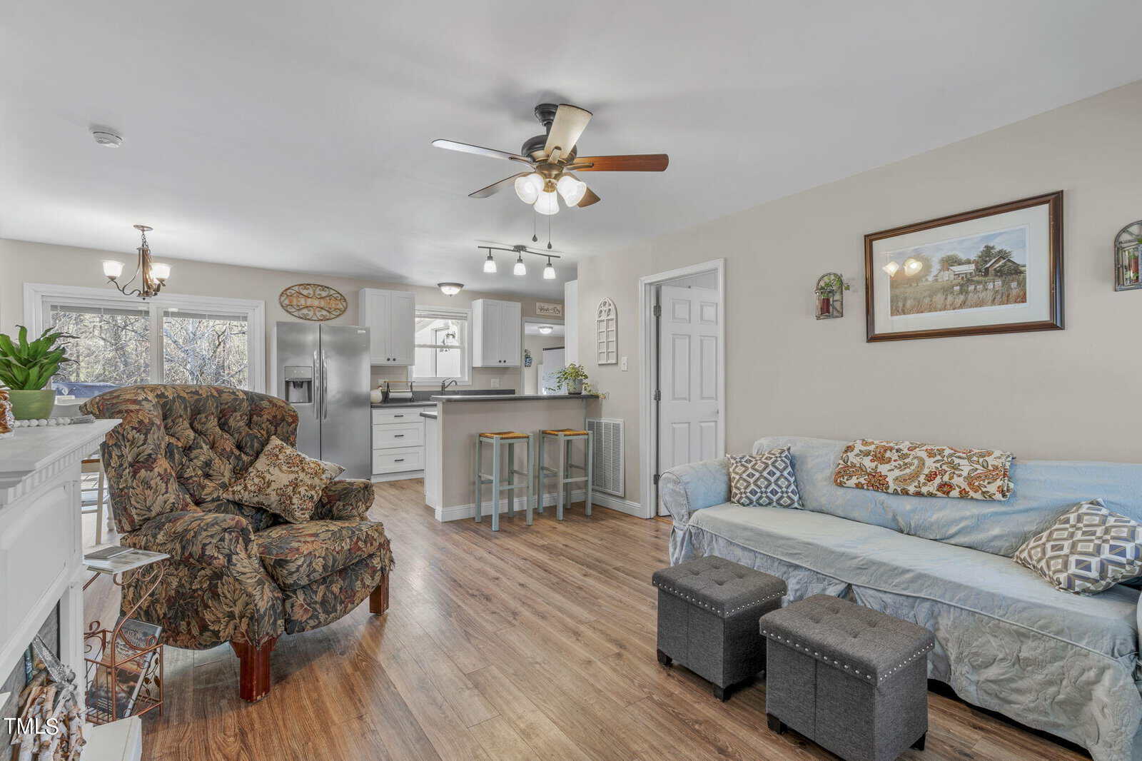 A cozy living room with a floral armchair, a blue sofa, two ottomans, and a fireplace. The space leads to a modern kitchen with bar stools and pendant lights. Natural light streams in through the windows and the room has a ceiling fan.
