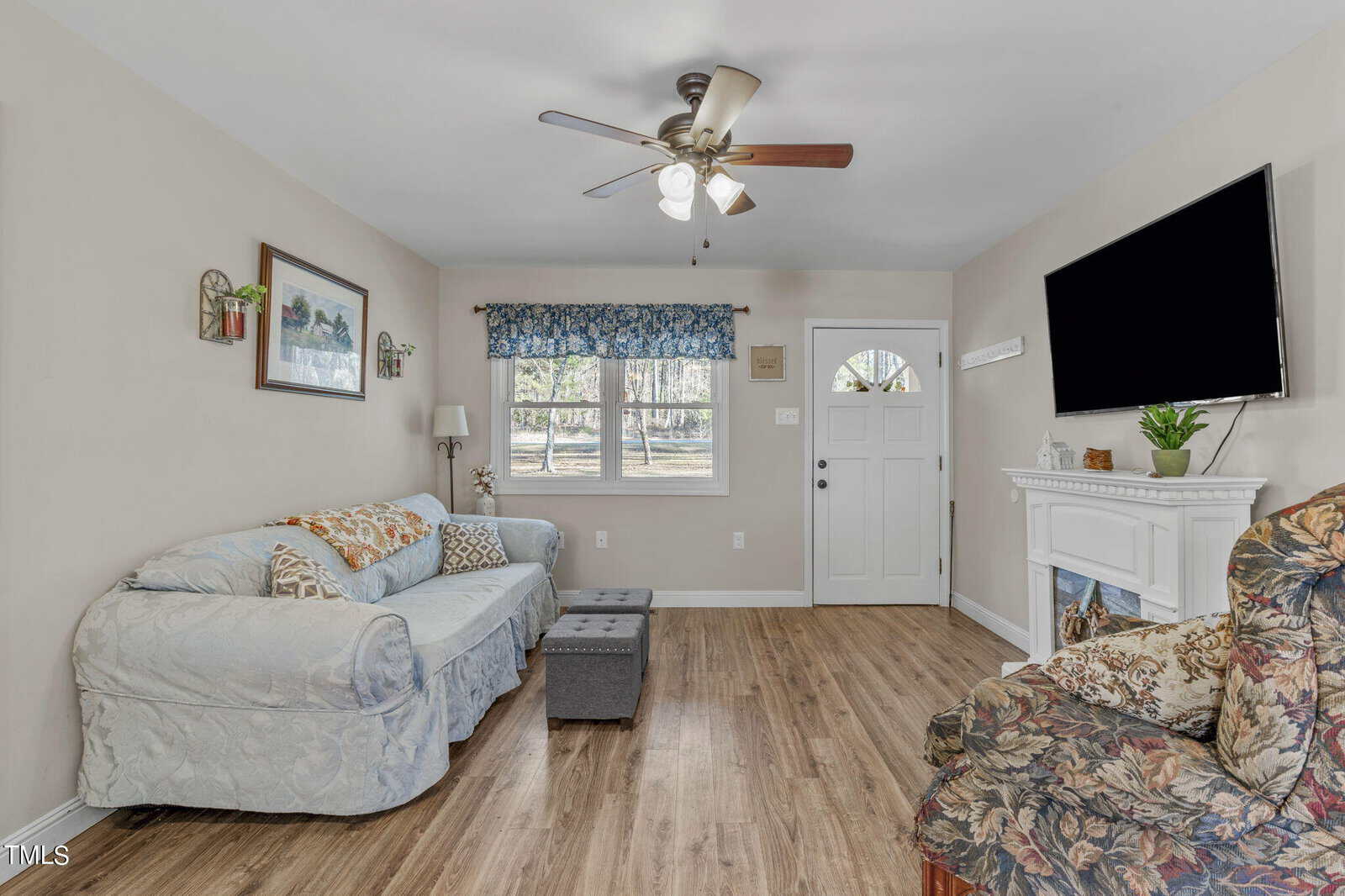 A cozy living room with a light gray sofa and patterned armchair. A flat-screen TV is mounted above a white fireplace. The room has wooden flooring, a ceiling fan, and a window with floral curtains. A small ottoman and artwork complete the decor.