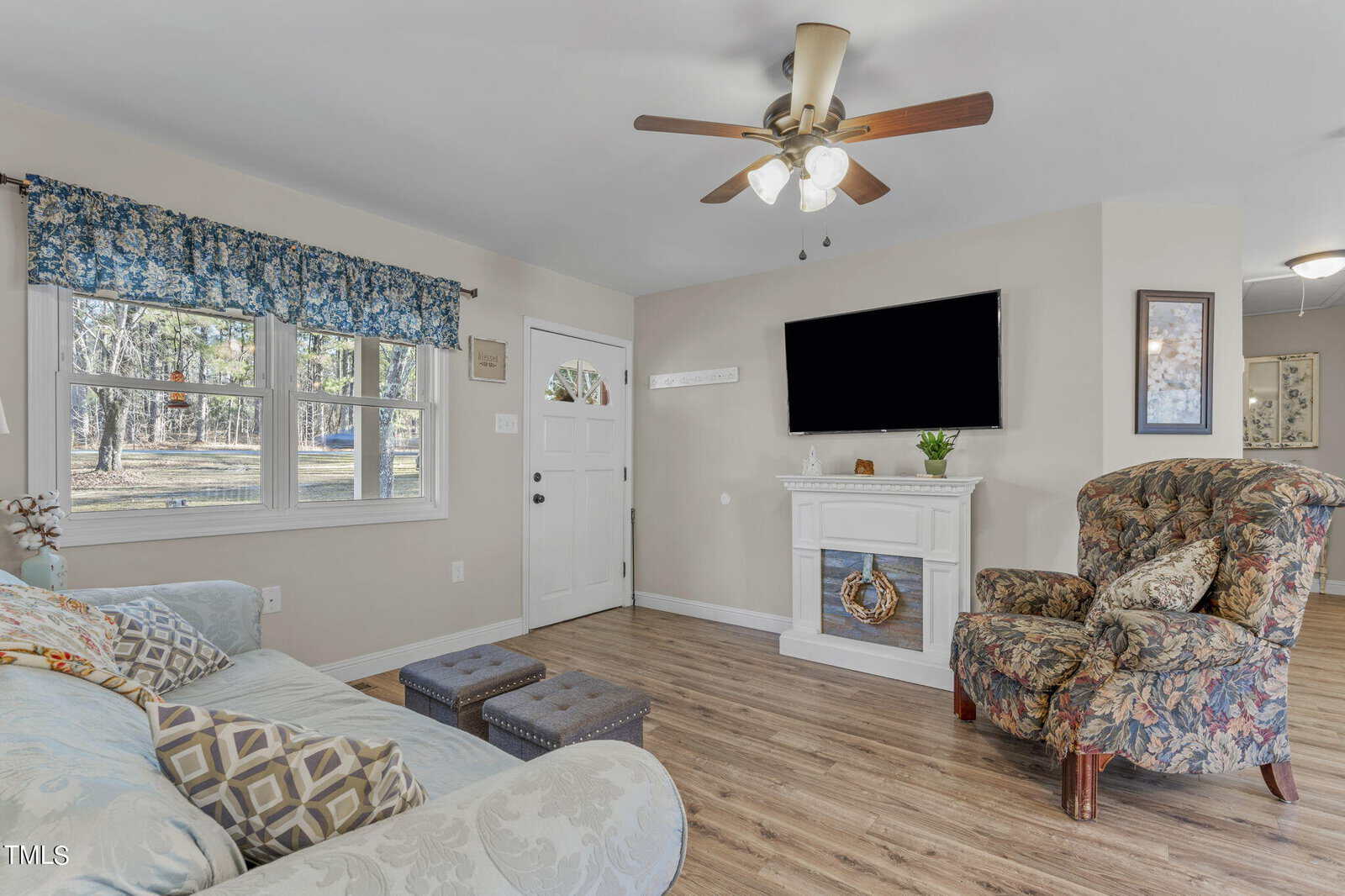 A cozy living room with a floral armchair, light blue sofa, and two small ottomans. A wall-mounted TV is above a white fireplace adorned with a decorative wreath. A ceiling fan is overhead, and large windows offer a view of the trees outside.