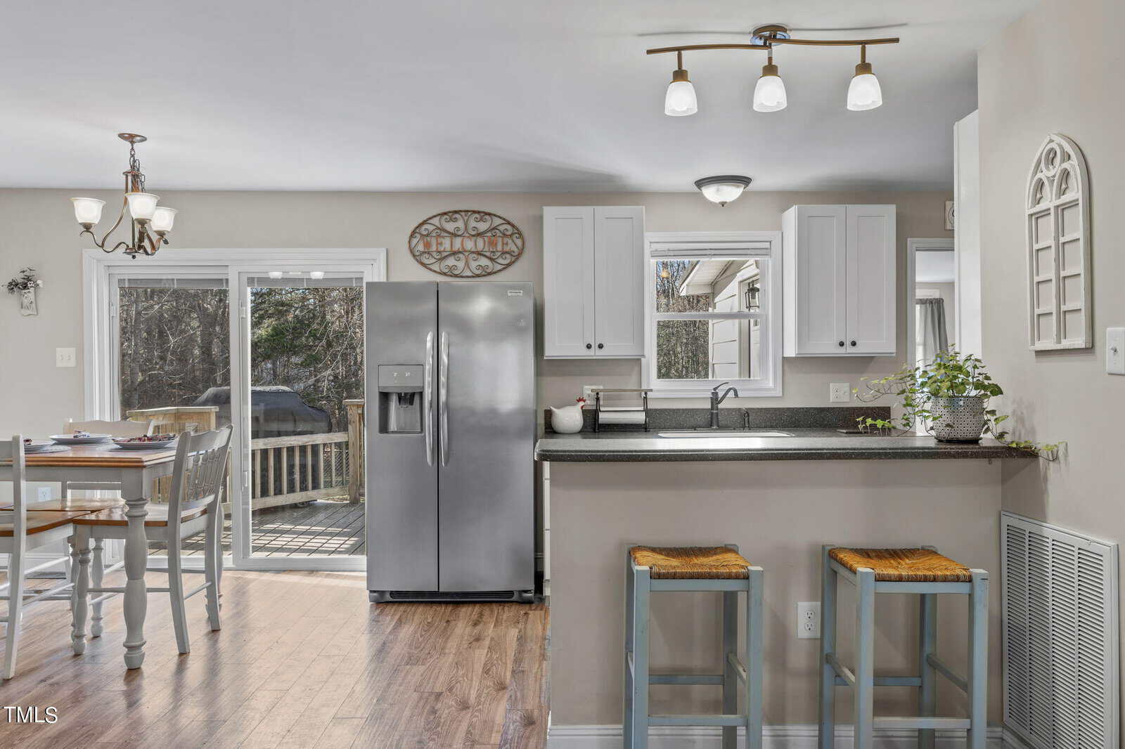 A bright kitchen with white cabinets, a stainless steel refrigerator, and a granite countertop bar with two stools. A dining table is near glass doors that lead to a deck. A decorative wall piece and small potted plant are visible.