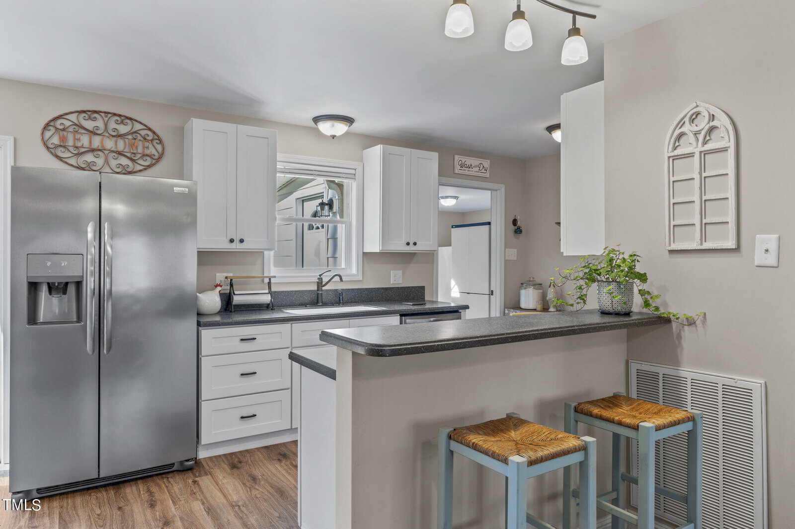 A modern kitchen featuring white cabinets, a stainless steel refrigerator, and a dark countertop. Two stools are tucked under the counter, and a decorative plant sits on the surface. The floor is wood, and ceiling lights are illuminated.