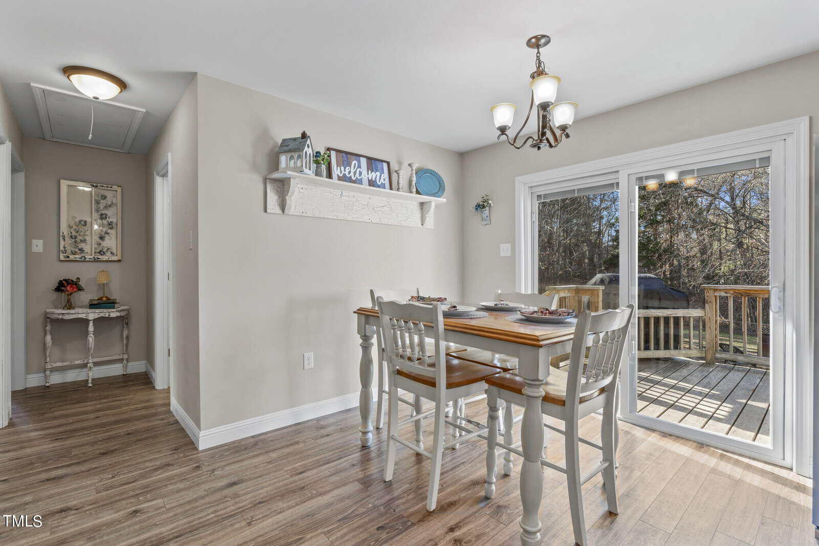 Dining room with wooden table and four chairs, set for a meal. Light beige walls with a decorative shelf and a "welcome" sign. Large glass doors open to a wooden deck with a view of trees. Hardwood floors and modern light fixture overhead.